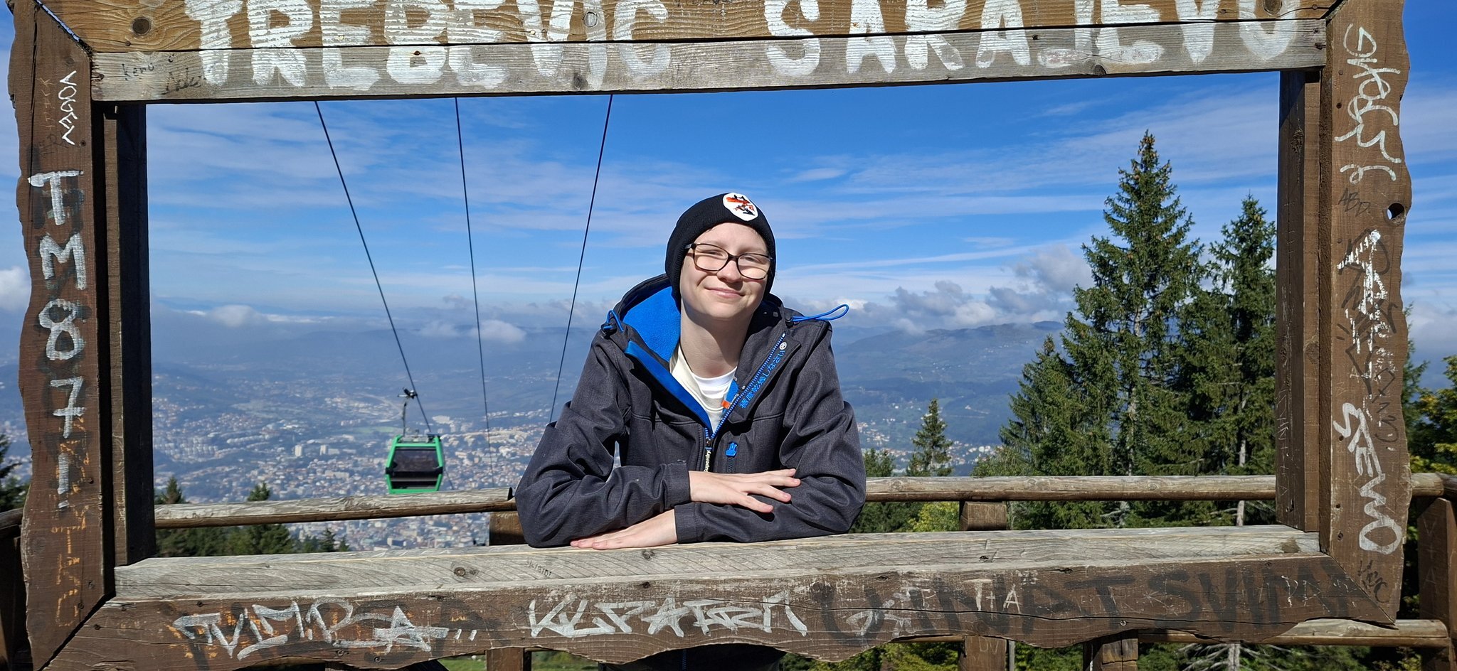 Picture of Enis in Sarajevo on the mountain behind the wooden frame with written white text: "Trebević Sarajevo" 