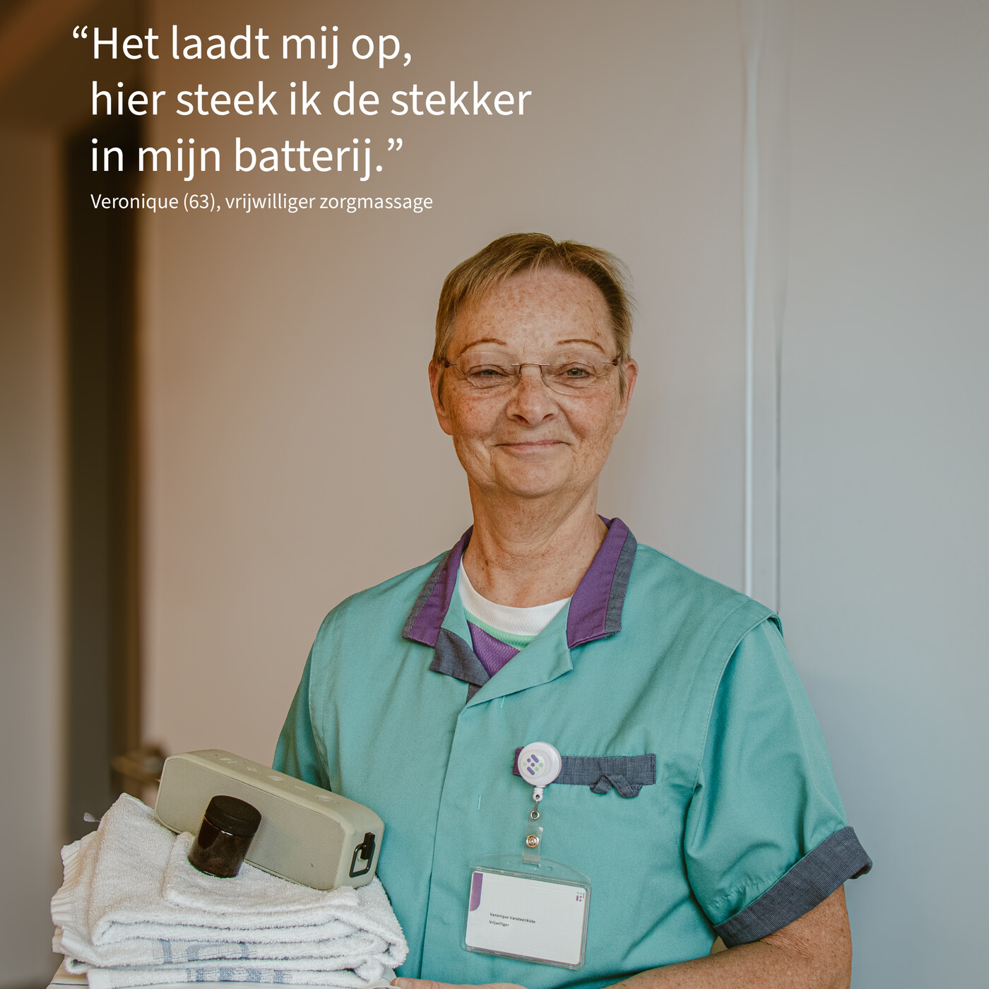 Portret van Veronique, vrijwilliger zorgmassage. Ze kijkt trots en draagt alles wat ze nodig heeft voor een massage in haar twee handen: handdoeken, een muziekbox en een flesje etherische olie.