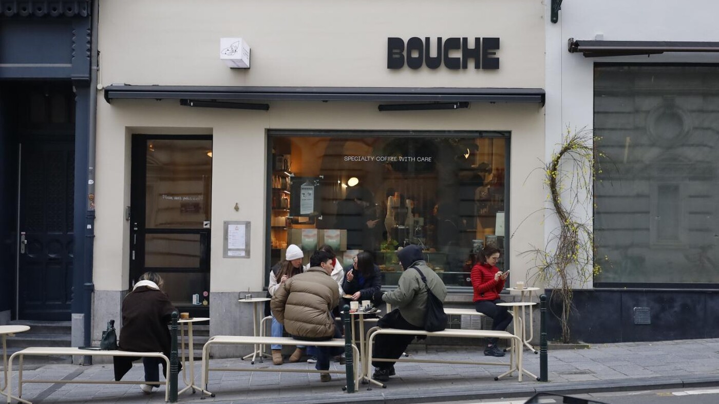 Street view of a café called “BOUCHE” in Brussels, with people sitting at outdoor benches drinking coffee and talking, while the warm interior is visible through a large front window.