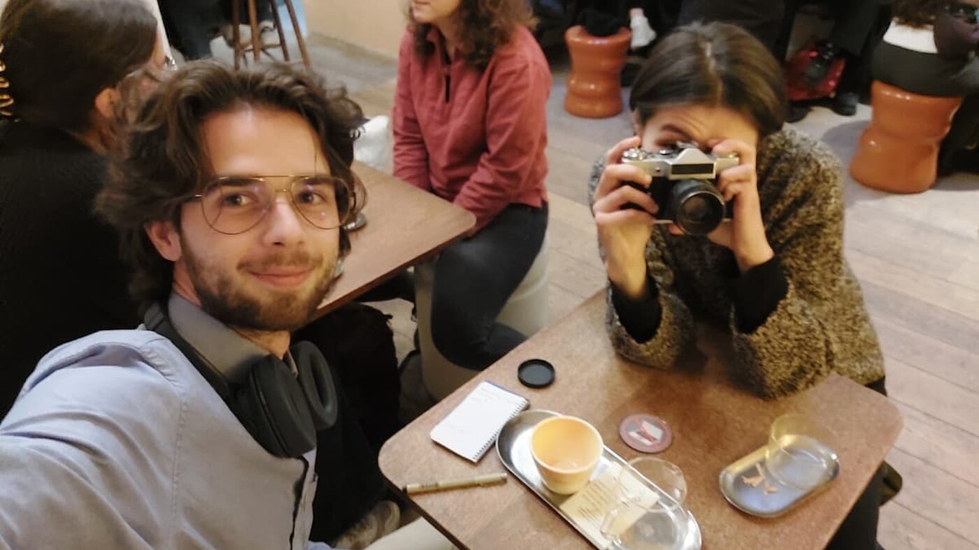 A man takes a selfie at a small café table while a woman across from him points an analog camera toward the lens; people sit and talk at nearby tables in a lively indoor setting.