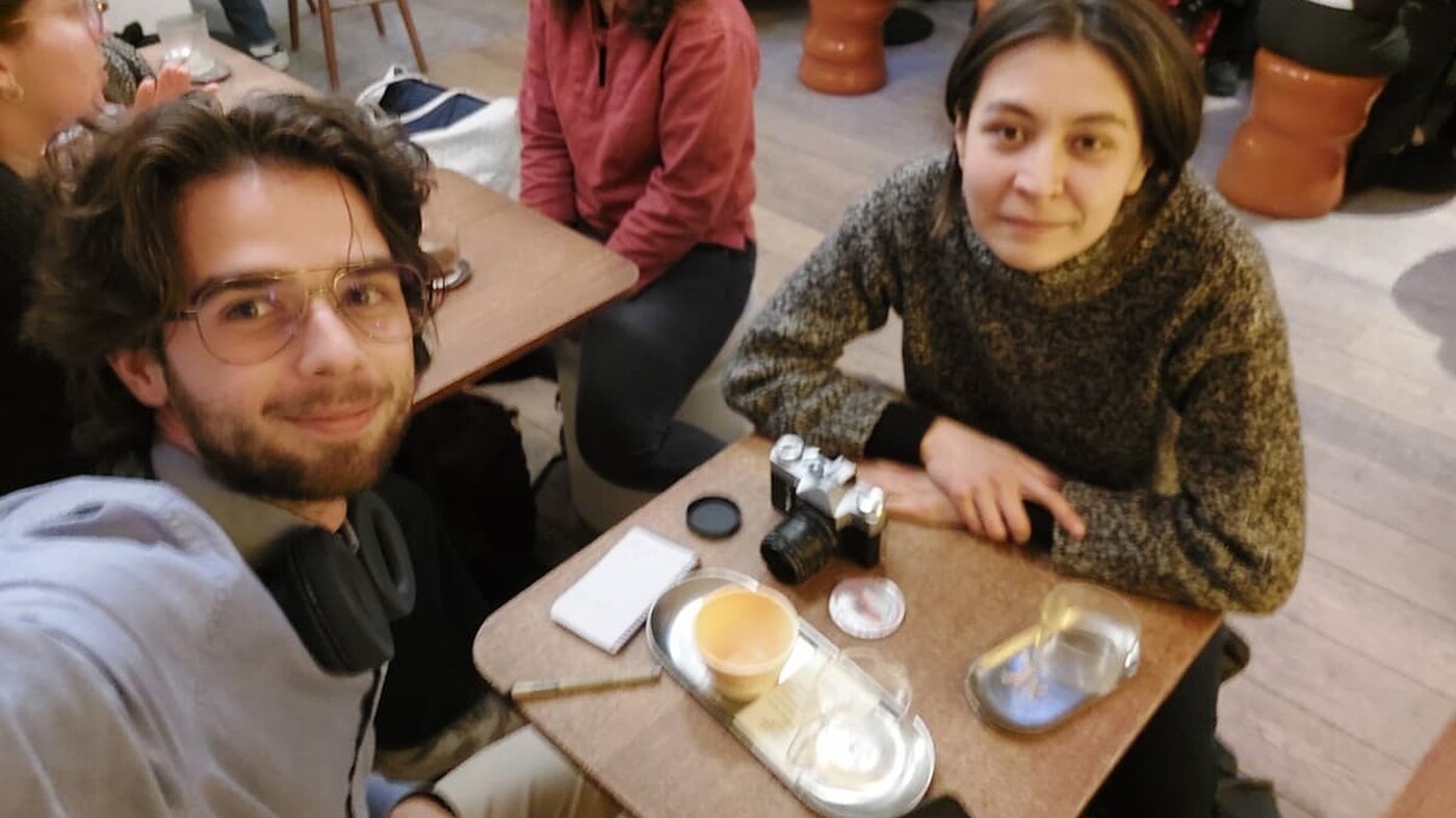 A man takes a selfie at a small café table while a woman sits across from him, looking at the camera; a film camera, notebook, and empty cups are on the table, with other people chatting in the background.